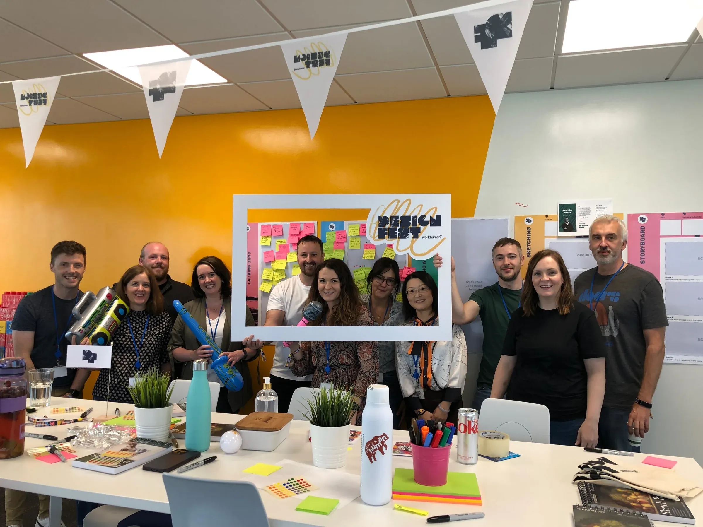 A team standing behind a table of sticky notes, holding a Design Fest frame