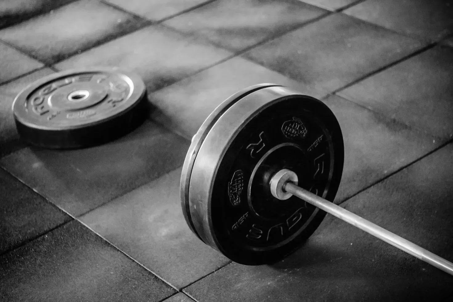 A black and white photograph of a loaded barbell resting on a rubber gym floor, with a spare plate lying beside it
